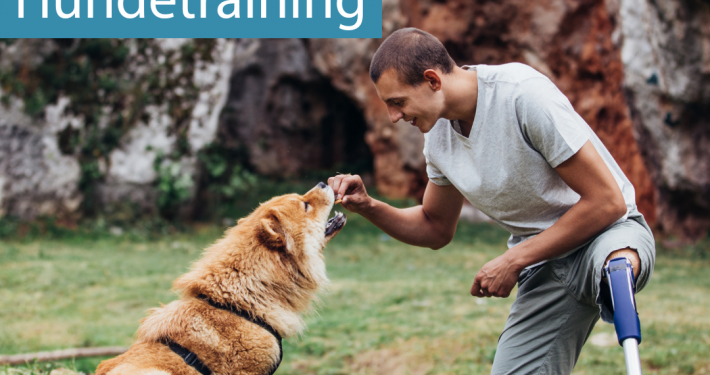 Ein junger Mann mit einer Beinprothese kniet auf einem Knie auf dem Boden und hält seinem Hund, einem Chow Chow ein Leckerchen vor die Nase. Der Hund sitzt auf der Wiese, i Hintergrund Natur, der Mann lächelt, beide sind entspannt und aufmerksam.